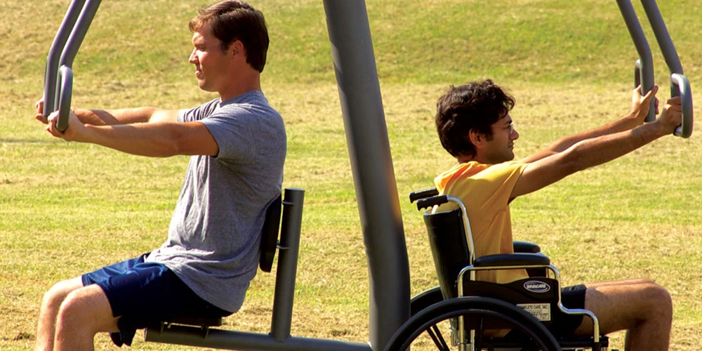 Man and wheelchair user exercising on accessible outdoor chest press machine in a public park fitness area